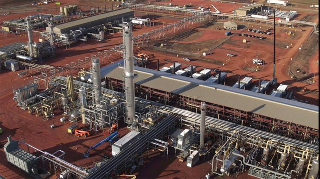 Aerial view of an industrial processing plant with multiple tall towers, pipelines, and surrounding red soil.