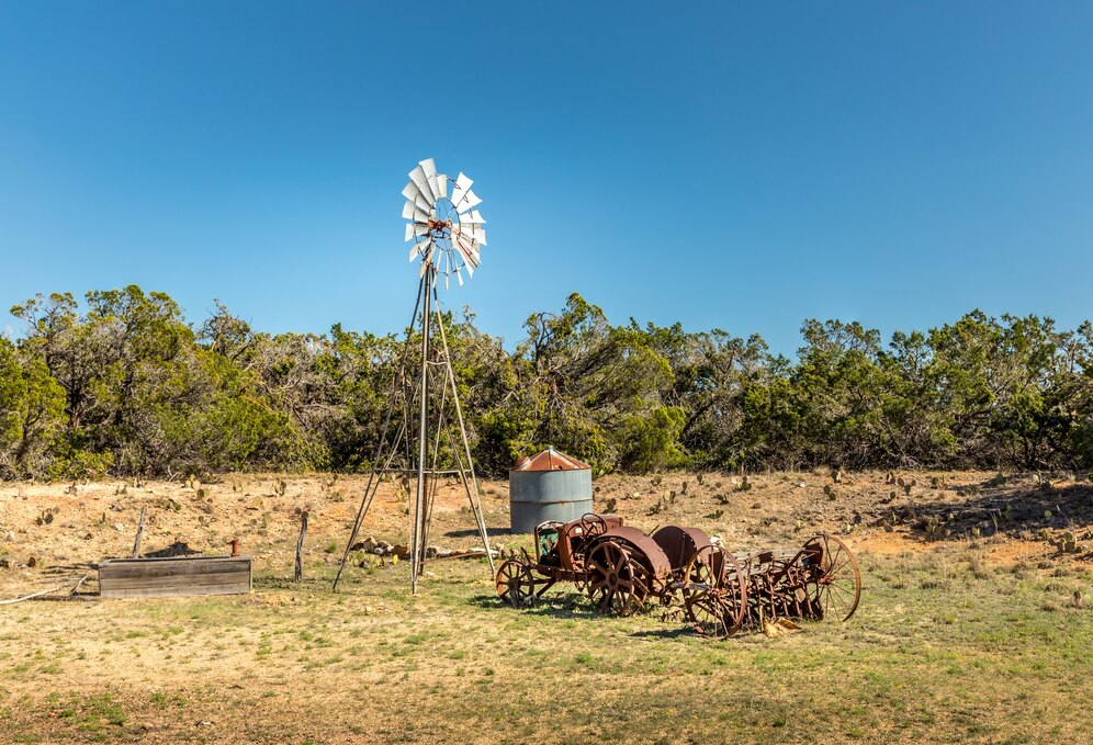 Rural Texas landscape featuring an old rusty tractor, a classic windmill, and dry grassy fields under a clear blue sky.