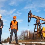 Two workers discussing renewable energy and oil extraction, with one holding a solar panel and the other wearing an orange safety vest near an oil pump jack.