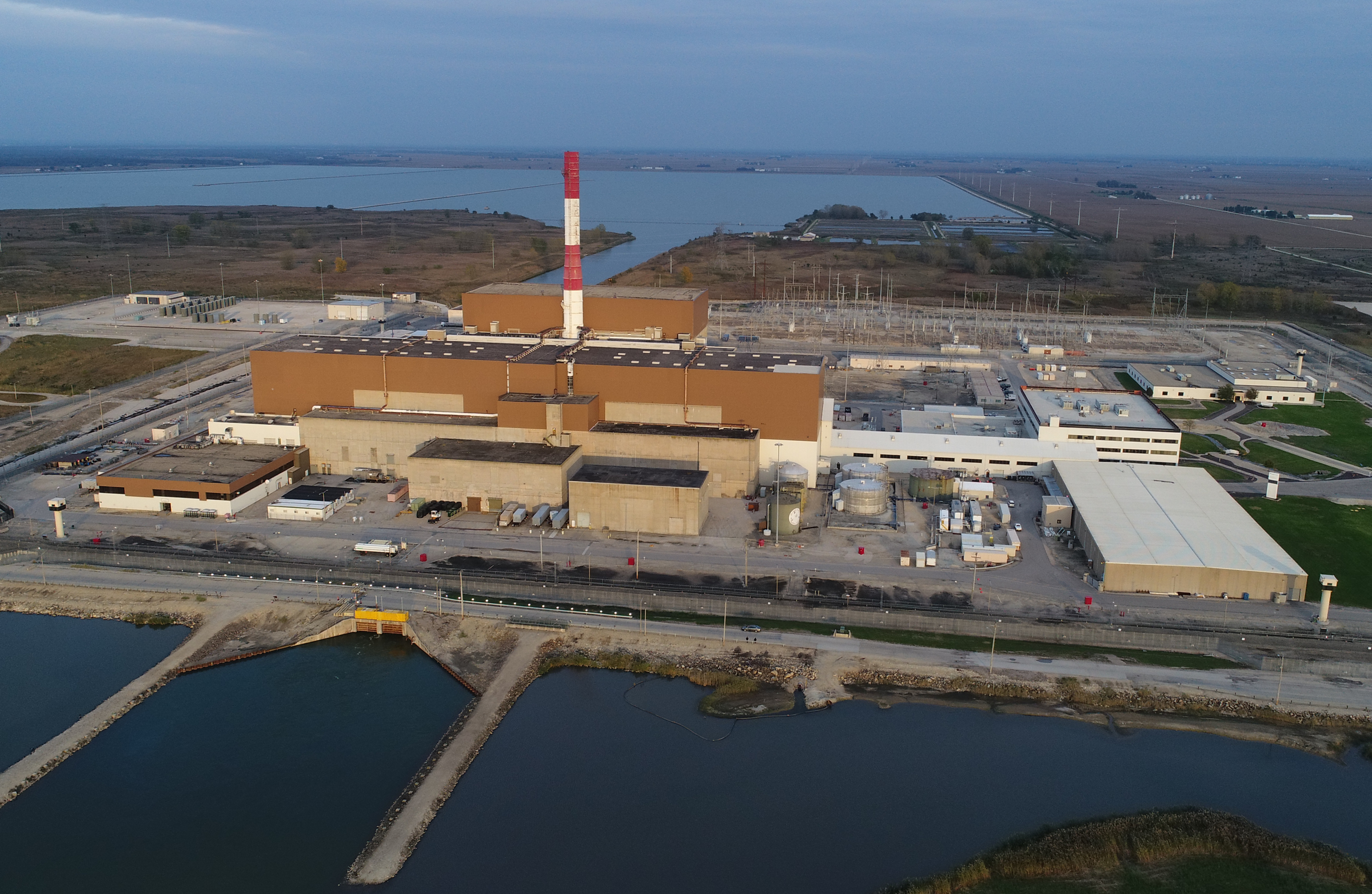 Aerial drone shot of a large industrial facility with a red-and-white smokestack near a water reservoir.
