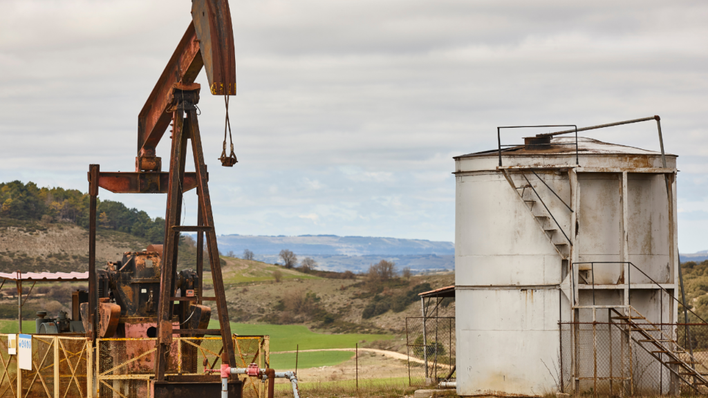 Rusty oil pumpjack and large storage tank in a remote oil field with rolling hills in the background.