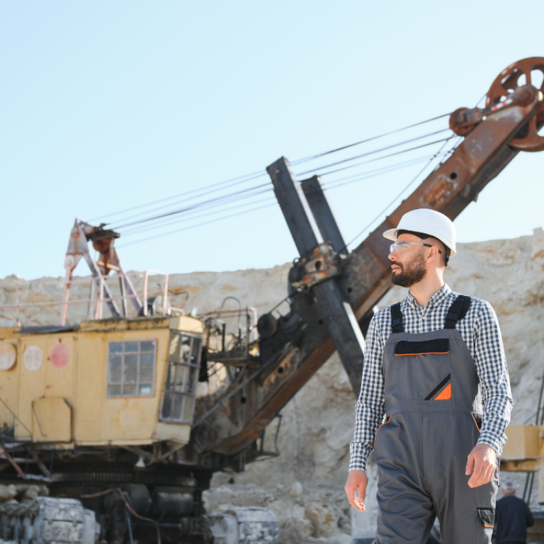 Worker in safety gear walking through an open-pit mining site with a large excavation machine in the background.
