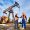 Two men wearing overalls, orange vests, and helmets, standing on oil field next to oil pump jack shaking hands, blue sky on background. Concept of business, petroleum industry and oil extraction.